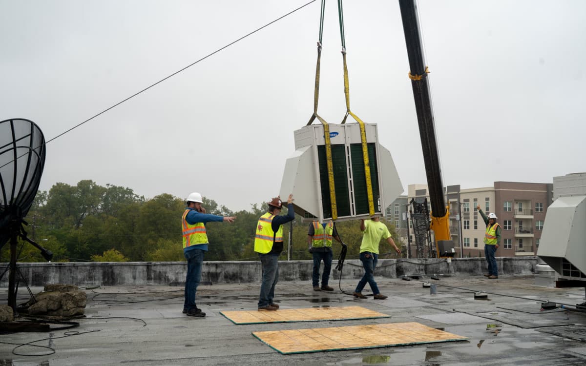 Crew of five workers guiding crane-lifted HVAC unit on commercial rooftop