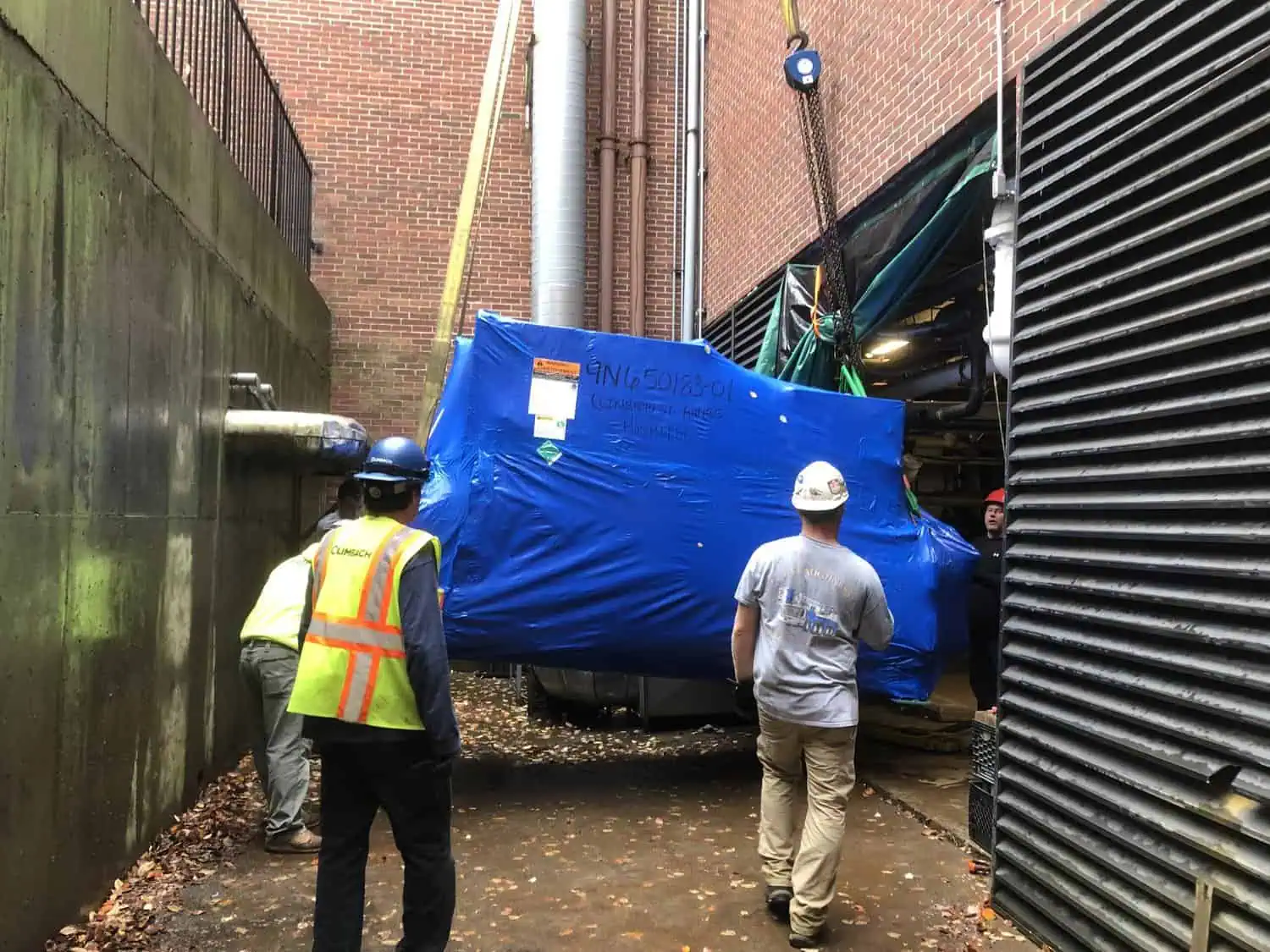 Crew of workers guiding crane-lifted blue-wrapped HVAC equipment between brick buildings