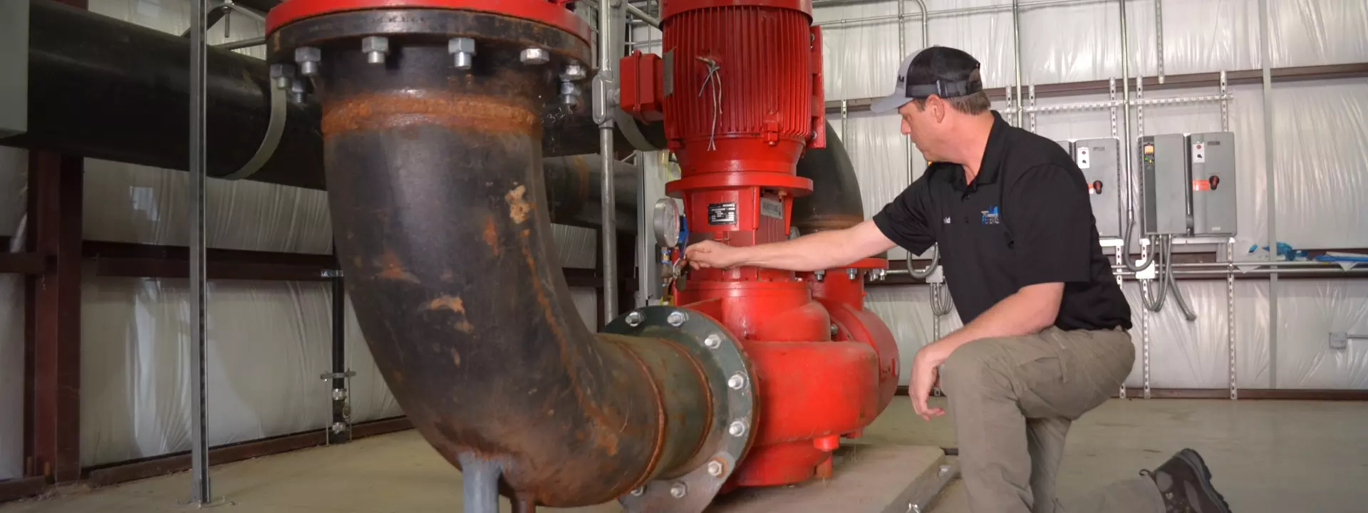 Technician kneeling beside large red industrial circulation pump checking gauge in mechanical room